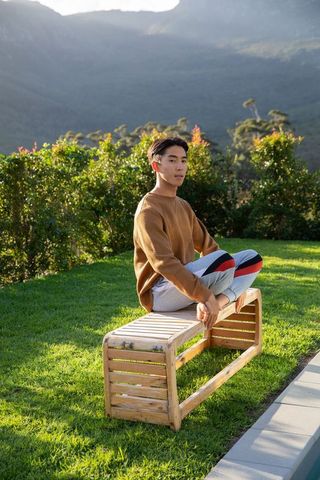 Young asian man sitting barefoot cross-legged on wooden bench by pool in mountain garden