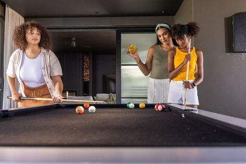Diverse women playing pool, holding cues and sipping drinks around black-felt table