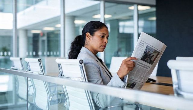 Executive woman reading newspaper in glass conference room, sitting at boardroom table
