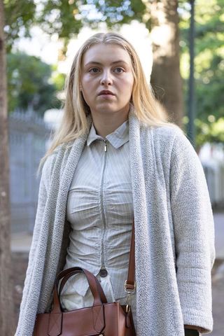 Young woman standing on tree-lined sidewalk in gray cardigan carrying brown leather tote