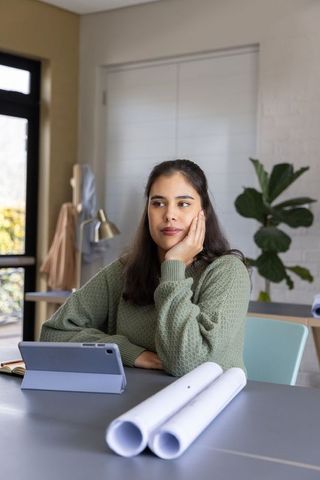 Contemplative asian woman planning with tablet and architectural blueprints at home office