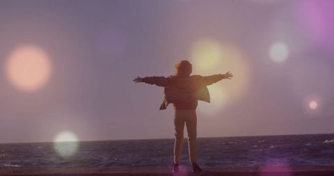 Woman standing with arms outstretched facing ocean at dusk with lens flare and serene bokeh