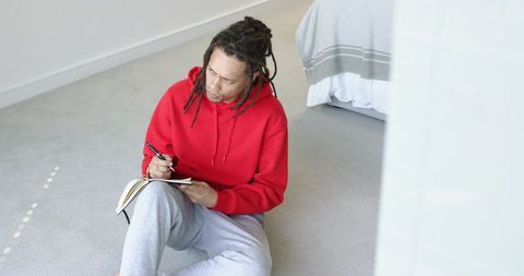 Mid 30s man with dreadlocks writing in notebook on bedroom floor in red hoodie, cozy soft light