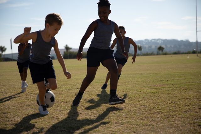 Multiracial elementary schoolboys are playing soccer on a school field during a sunny day. This image can be used for promoting physical education, team sports, childhood activities, and outdoor play. It is ideal for educational materials, sports training programs, and advertisements for children's sports equipment.