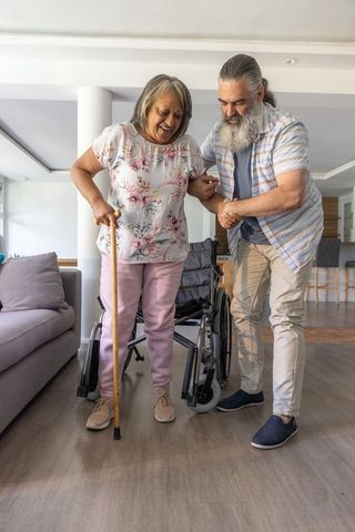 Senior woman rising from wheelchair using cane while partner assisting at home