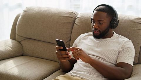 Man relaxing on beige sofa wearing striped tee listening with headphones using smartphone