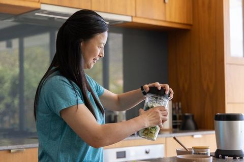 Mature asian woman making green smoothie using personal blender in sunlit modern kitchen