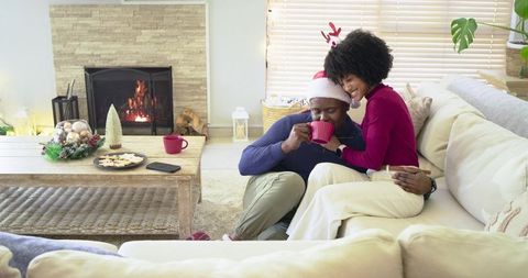 Diverse couple cuddling on sofa wearing Santa hat sipping red mug by cozy fireplace