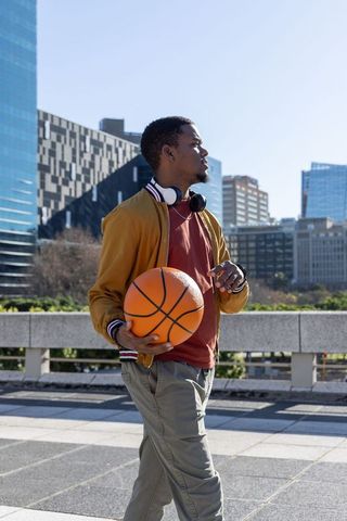 African american man walking urban plaza holding basketball wearing headphones