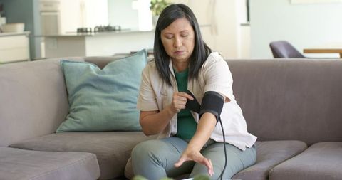 Woman measuring blood pressure at home on couch using arm cuff in calm modern living room