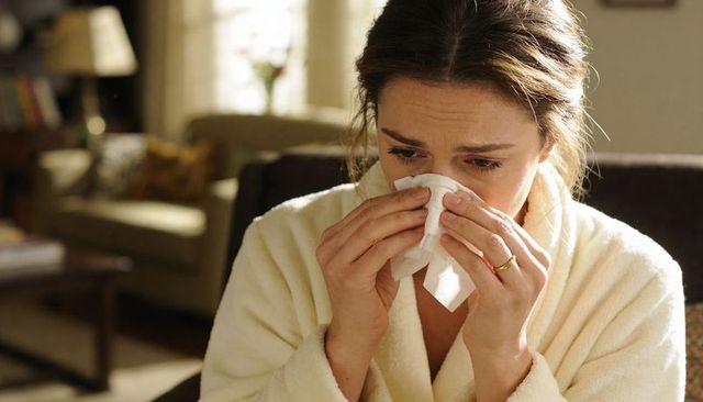 Woman blowing nose in bathrobe at home conveying illness and emotional fatigue