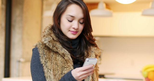 This image features a woman in a casual fur vest engaging with her smartphone in a stylish modern home setting. Ideal for use in lifestyle blogs, technology-related articles, and advertisements focusing on casual fashion or home living.