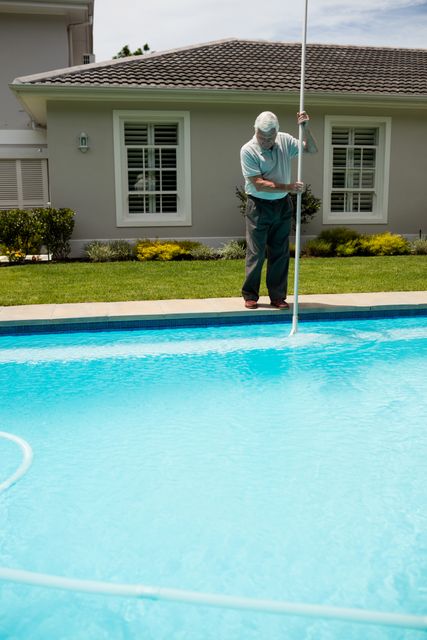 Senior man cleaning swimming pool with a long pole on a sunny day. Ideal for illustrating themes of outdoor maintenance, elderly activities, summer leisure, and residential care. Useful for articles, advertisements, and blogs related to home care, retirement living, and pool maintenance services.