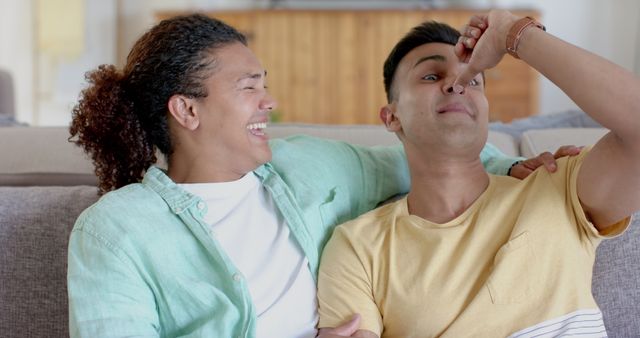 Happy scene showing an African American man pinching the nose of his embraced friend as they sit on a couch, laughing in a living room setting. Perfect for depicting themes of friendship, bonding, and domestic life. Useful for promoting casual and cheerful home environments, highlighting diverse friendships, and creating feel-good content.
