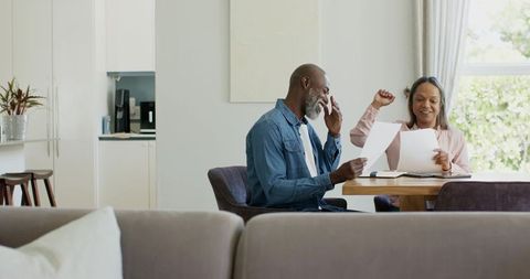 Diverse mature couple reviewing documents at home dining table celebrating good news