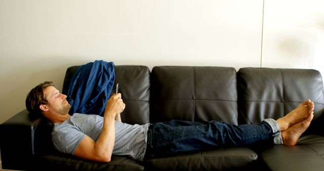 Man lying on black leather sofa, using smartphone in a casual home environment. He is wearing jeans and a t-shirt, creating a relaxed and comfortable vibe. Ideal for illustrating concepts of technology use at home, relaxation, leisure activities, modern lifestyle, and casual attire.