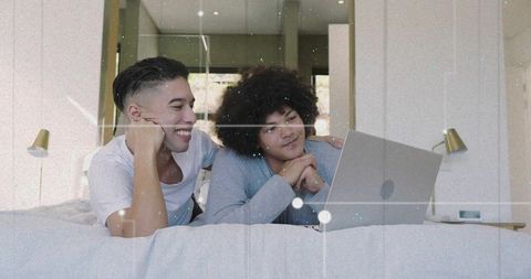 Two young men relaxing on bed using laptop, sharing cozy home browsing and connection