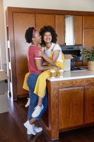 Laughing african american female couple unpacking in new kitchen sharing coffee