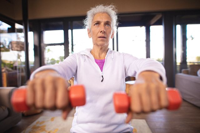 Senior woman in white hoodie lifting dumbbells at home. Promotes active aging, healthy lifestyle, and indoor fitness. Can be used in content about senior fitness, home workouts, and retirement life.