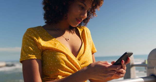 Focused biracial woman in yellow dress using smartphone on sunny promenade by the sea. Communication, summer, vacations, lifestyle and free time, unaltered.