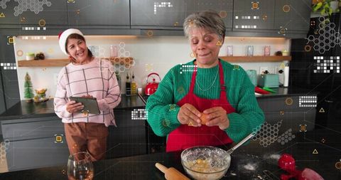 Senior woman cracking egg while baking festive holiday cookies with friend using tablet
