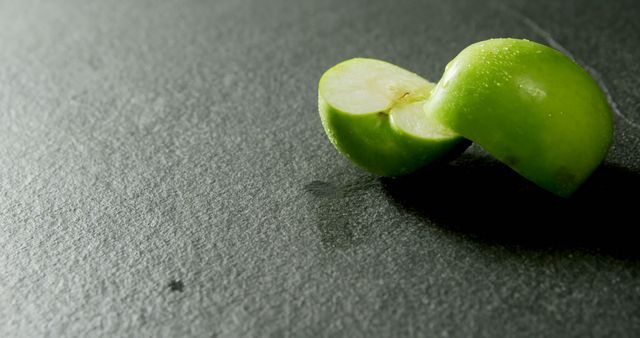 Sliced green apple resting on a dark slate surface. This close-up captures the freshness and juiciness of the apple, making it perfect for promoting healthy eating, nutrition, or natural food products. It can also be used in advertisements for organic foods, diet plans, or as a simple yet elegant addition to a health and wellness blog.