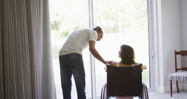 Biracial woman in wheelchair and smiling male partner talking and looking out of living room window. wellbeing and domestic lifestyle with physical disability.