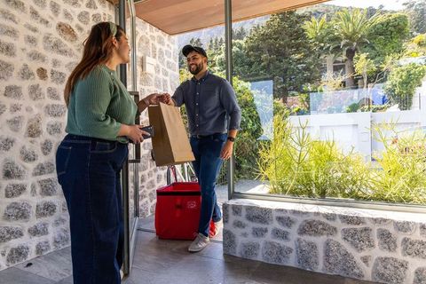 Delivery driver handing grocery bag to asian woman at home entrance, smiling