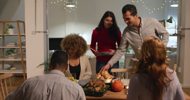 Front view of a group of young multi-ethnic male and female friends bringing dishes of food and sitting down at a table for a roast turkey dinner at home for Thanksgiving