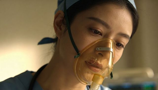 Asian nurse wearing oxygen mask providing compassionate bedside care hospital ward close-up