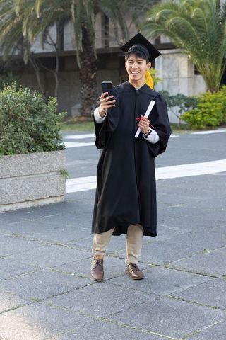 Asian graduate wearing cap and gown holding diploma and taking selfie on university campus