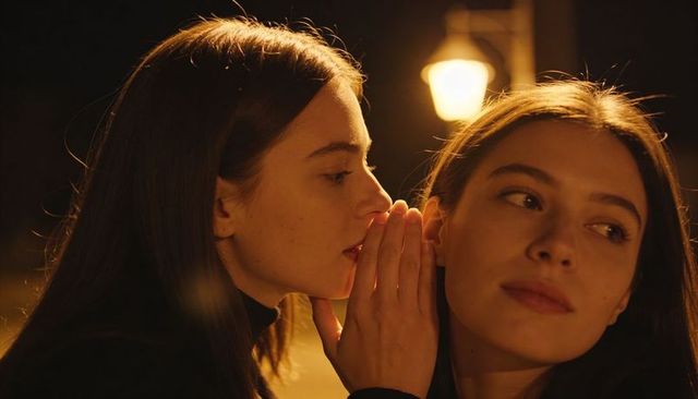 Whispering secret on night street, young women leaning close under warm lamppost glow, intimate