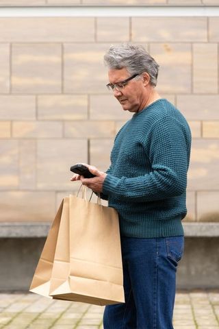 Senior man checking smartphone while carrying brown paper shopping bags by stone facade