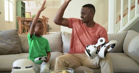 African American father and son high-fiving while playing robot toy on sunlit sofa