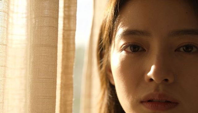 Asian woman gazing through sheer beige curtain, warm window light, contemplative portrait