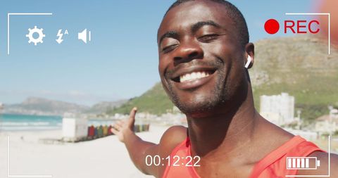 Young African American man smiling and recording a video on a beach. He appears joyful and healthy, suggesting a modern and active lifestyle. This visual can be used to promote outdoor fitness activities, healthy living campaigns, or travel blogs highlighting vibrant coastal experiences.