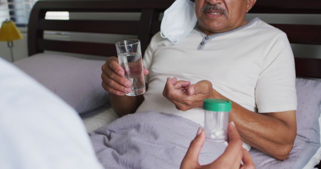 Elderly man sitting in bed holding a glass of water with one hand and medication in the other, assisted by healthcare professional holding a medicine container. Perfect for illustrating elderly care, home health services, patient support, and senior medical treatment in promotional materials, brochures, and healthcare websites.