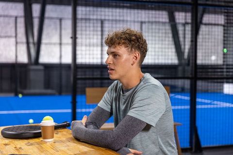Young man taking break after padel match holding coffee beside racket on indoor court