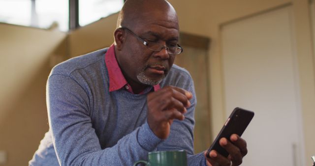 Mature African American man using smartphone in home setting. He is leaning on kitchen counter with a green mug. Ideal for content about lifestyle, technology use, communication, and senior demographics engaging with modern technology.