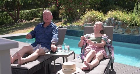 Asian senior couple relaxing on poolside loungers in sunny backyard retreat