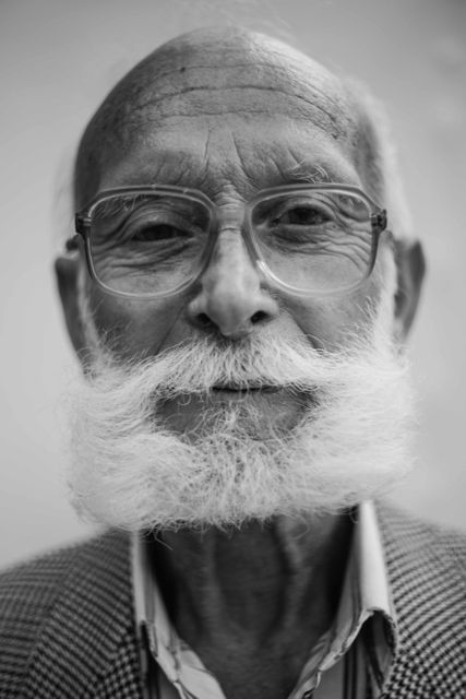 Close-up black and white portrait of an elderly gentleman with a distinctive white beard and glasses. The man's expressive face and prominent facial wrinkles highlight a lifetime of experiences and wisdom. Useful for articles focusing on aging, wisdom, life stories, and senior living topics. Ideal for advertisements and publications targeting senior audiences.