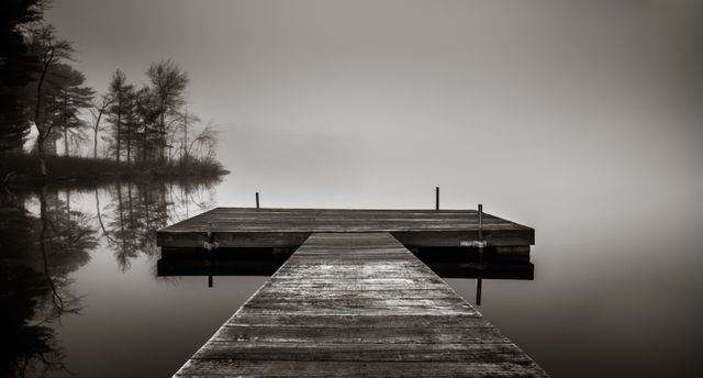 Wooden pier extending over a calm lake in morning mist. Surrounded by calm waters and faint, silhouetted trees. Ideal for themes related to tranquility, nature, meditation, and isolated retreats. Perfect for use in digital backgrounds, websites, or print media that focus on relaxation and serenity.
