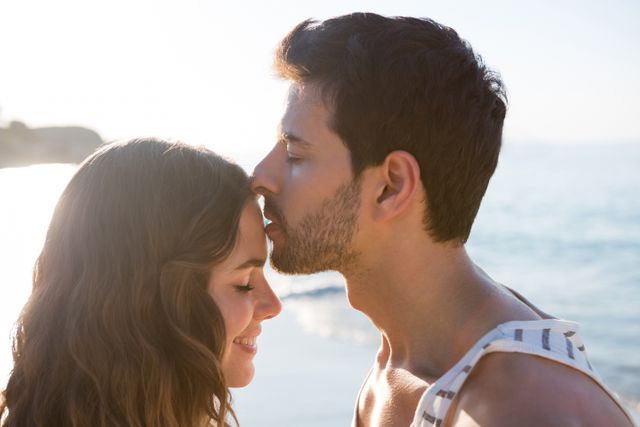 Side view of young man kissing girlfriend forehead at beach during sunny day