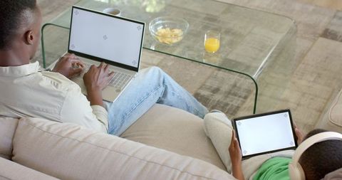 African american father and son using laptop and tablet on sofa wearing headphones