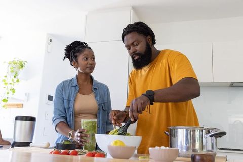 African couple preparing healthy meal, tossing salad and blending smoothie in bright kitchen