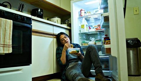 Asian cook sitting on kitchen floor eating takeout from open refrigerator at night
