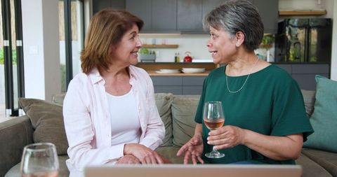 Hispanic female friends chatting in modern lounge with wine and laptop