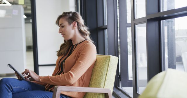 Young woman using tablet seated in a modern office lounge area. The setting is casual with natural light coming through large windows. Perfect for themes related to business, technology, modern work environments, digital connectivity, and professional settings. Ideal for corporate websites, office-related articles, and digital workspace promotions.