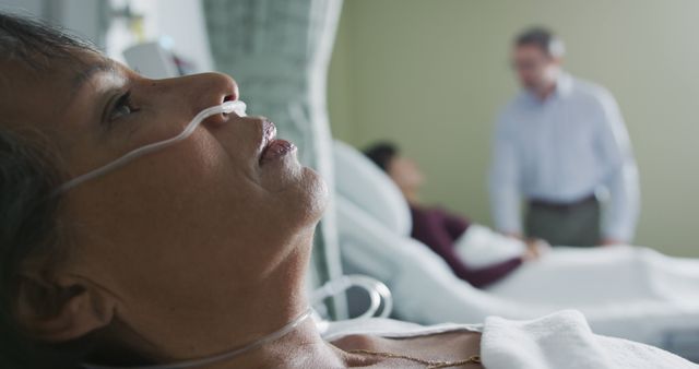 Patient is lying on a hospital bed with medical equipment attached. Two supportive visitors can be seen in the background. Useful for depicting healthcare situations, patient care, hospital visits, and emotional support scenes.