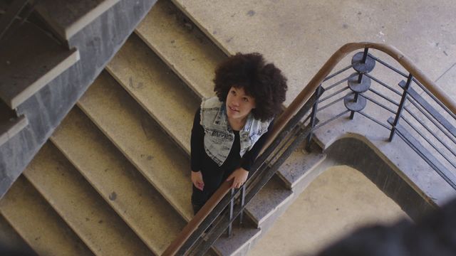 Woman with afro hairstyle climbing a staircase in an empty building. Denim vest adds a stylish touch. Video can be used for concepts of fashion, solitude, perspective, and architectural design in urban environments.